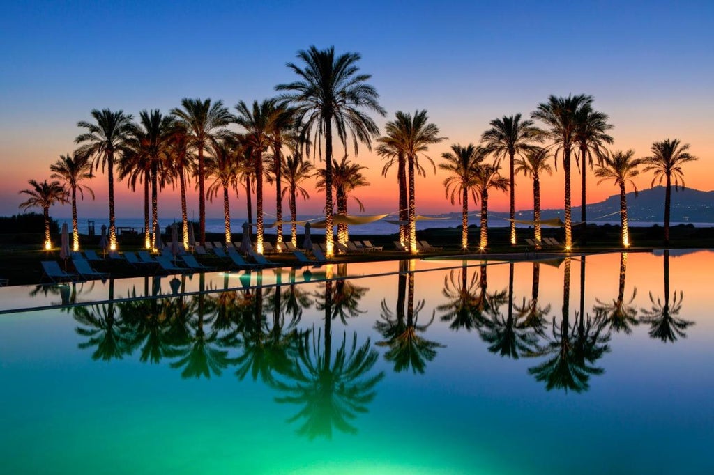 Elegant Mediterranean beachfront resort with infinity pool overlooking Sicily coast, palm trees and cream buildings at golden hour