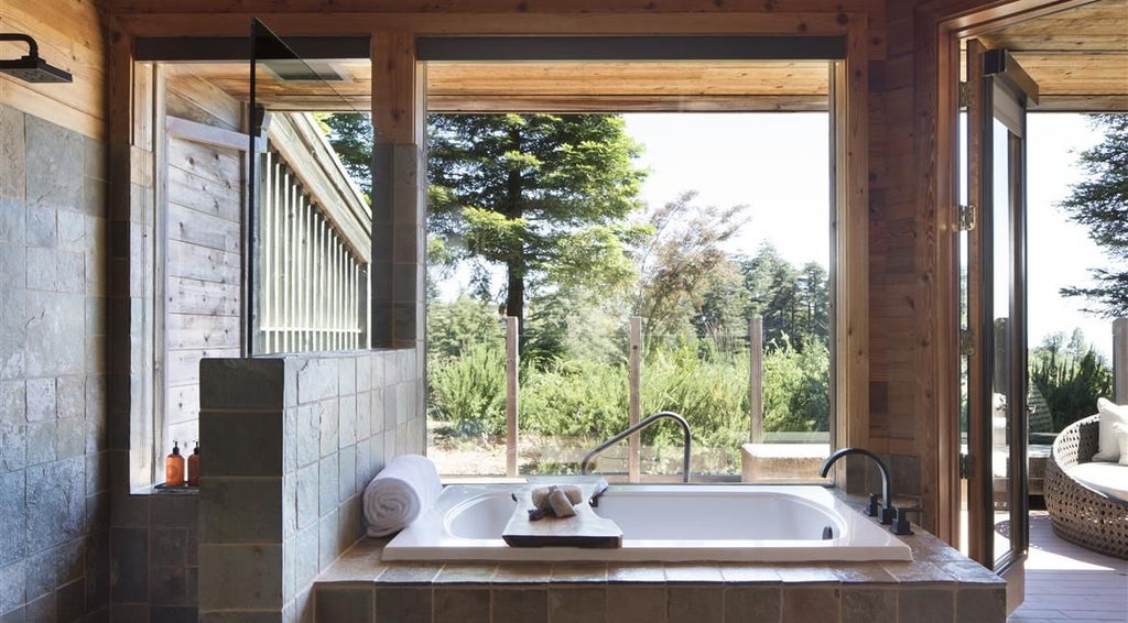 Luxurious hot tub suite overlooking rugged coastline, with modern wood-paneled interior, plush furnishings, and expansive Pacific Ocean panoramic view through floor-to-ceiling windows