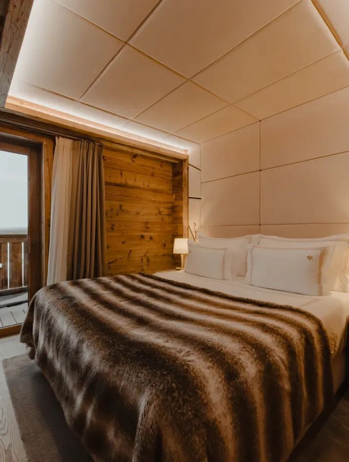 Elegant alpine ski resort suite with modern minimalist design, warm wood tones, plush white bedding, panoramic mountain view windows, soft lighting