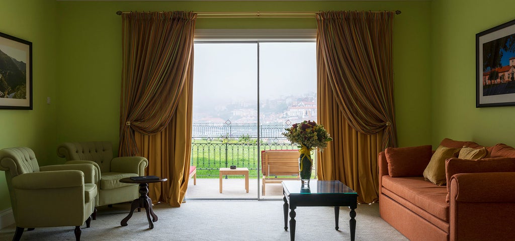 Elegant hotel suite with floor-to-ceiling windows, plush king bed, chic white furnishings, and stunning views of Porto's Douro River