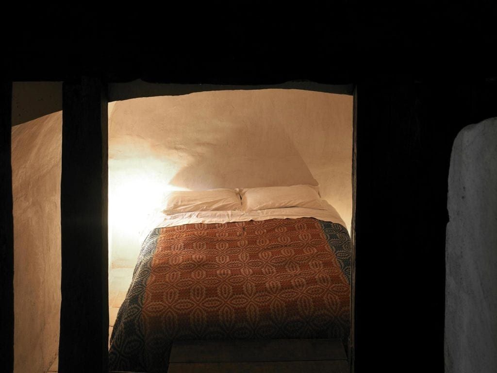 Rustic Italian hotel room with stone walls, arched window overlooking historic bell tower, featuring vintage wooden furnishings and soft, warm lighting