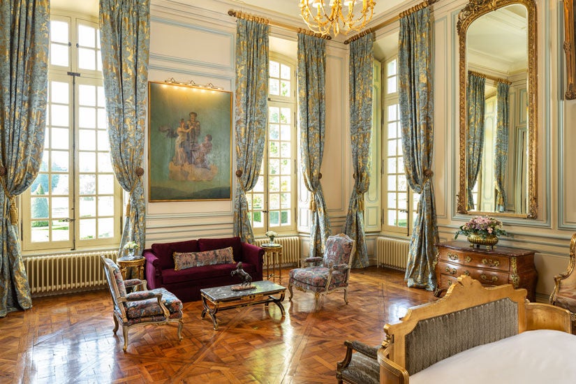 Opulent French château suite with ornate period furnishings, crystal chandelier, marble fireplace, and floor-to-ceiling windows with garden views