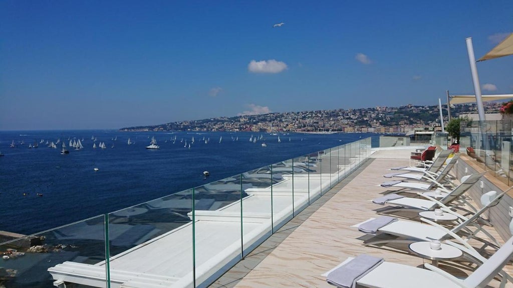 Elegant five-star hotel overlooking Naples Bay with white stone facade, ornate balconies, and Mediterranean gardens under blue sky