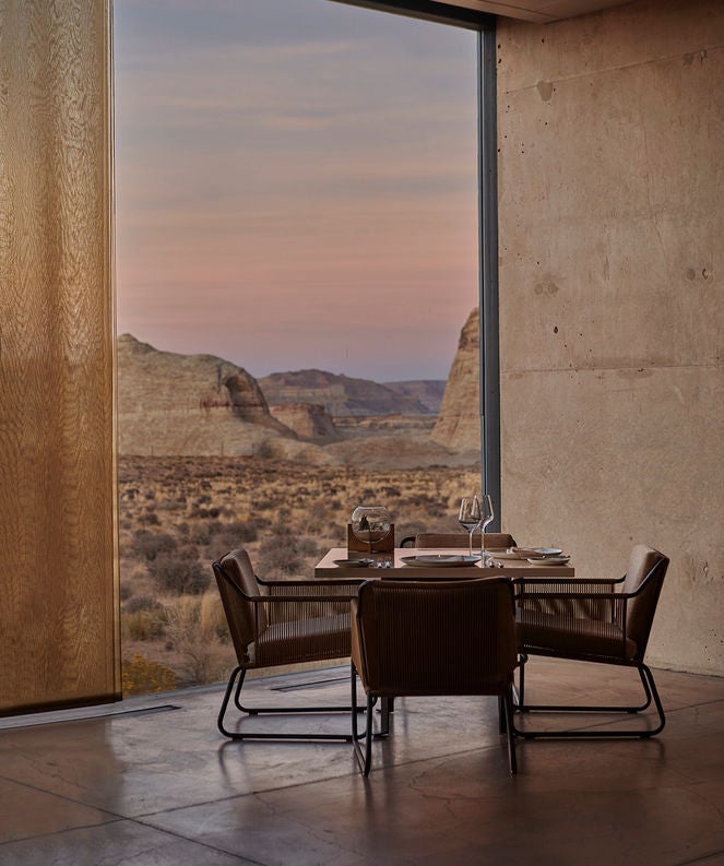 Modern luxury resort with geometric concrete buildings seamlessly integrated into desert landscape, framed by sandstone cliffs at sunset