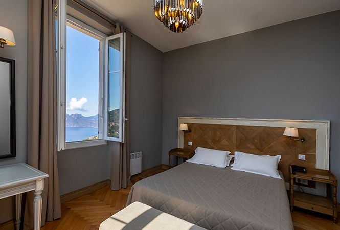 Minimalist seaside hotel room with white linens, blue Mediterranean views, and clean modern design featuring natural stone walls and large windows