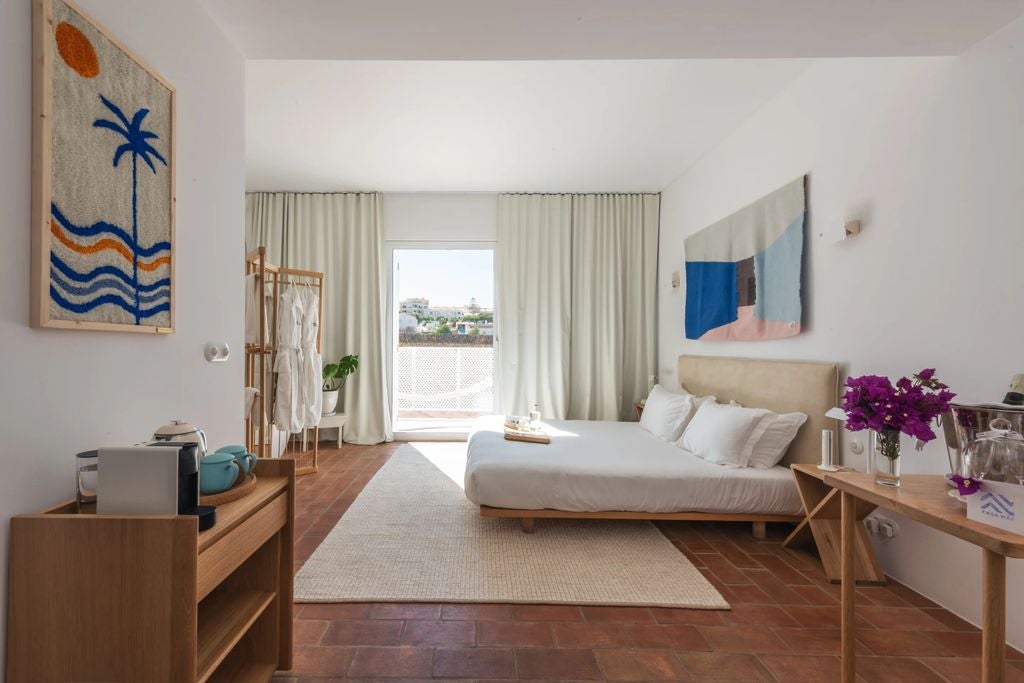 Modern minimalist hotel room with white walls, ornate wooden headboard, green potted plants, antique side table, and natural light from terrace doors