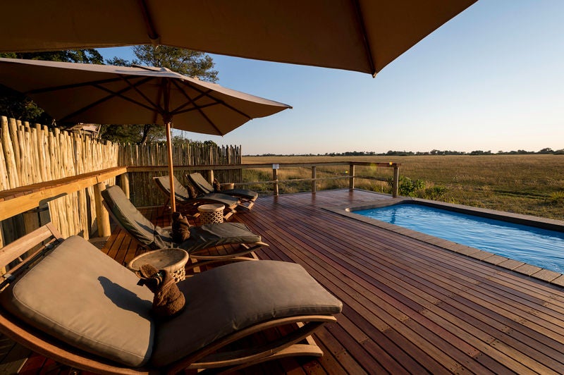 Elevated luxury safari lodge with wooden walkways connecting thatched suites overlooking vast Okavango Delta grasslands at sunset