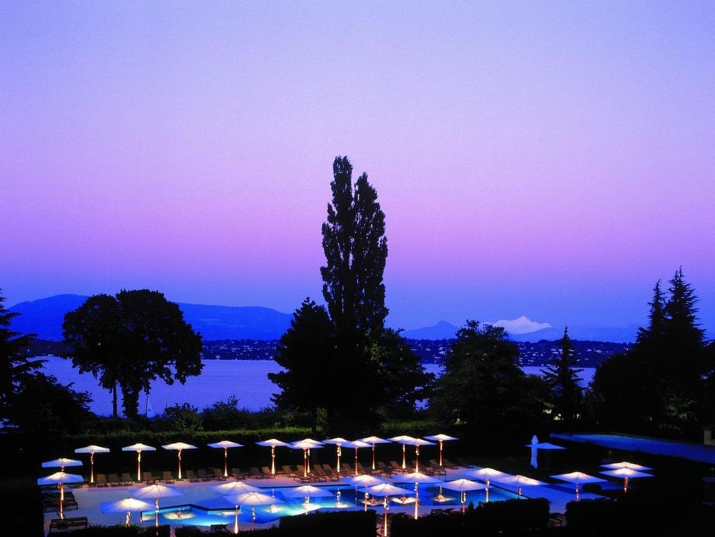 Elegant facade of La Reserve Geneva hotel reflecting in Lake Geneva, with manicured gardens and classic European architecture at sunset