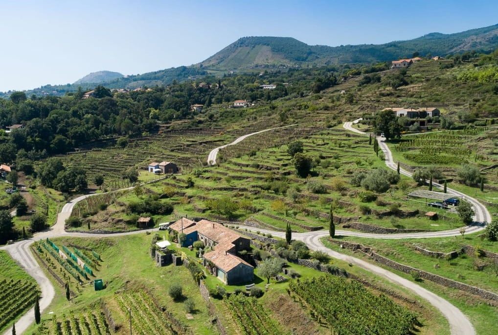 Elegant Sicilian boutique hotel nestled among lush vineyards, with renovated rustic stone farmhouse surrounded by verdant Mount Etna landscape at sunset