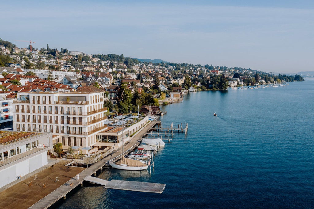 Contemporary lakeside hotel with floor-to-ceiling windows and private balconies reflecting on Lake Zurich, surrounded by Swiss Alps at sunset