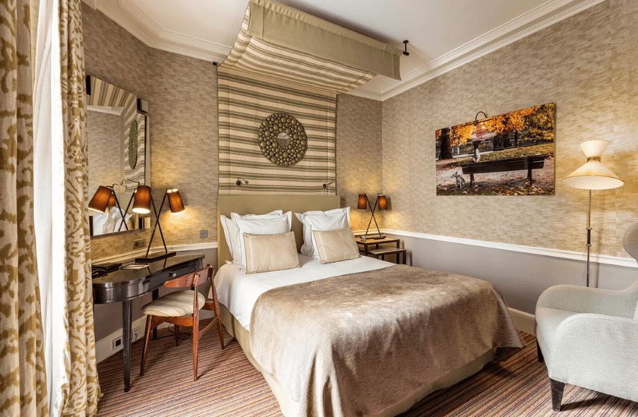 Elegant Parisian hotel room with soft neutral tones, plush king bed, ornate headboard, and classic French design elements creating a sophisticated ambiance