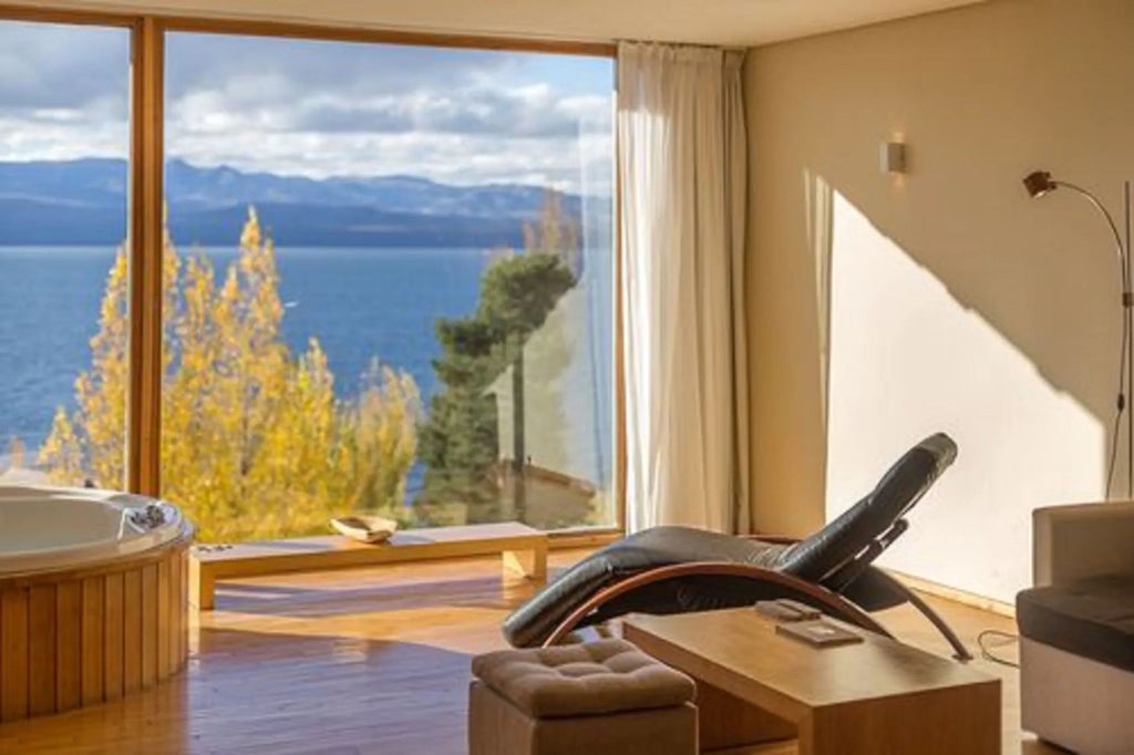 Modern lakefront hotel with glass facade, overlooking Nahuel Huapi Lake, framed by snow-capped Andes mountains and green forests