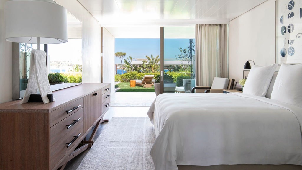 Elegant hotel room with floor-to-ceiling windows, private terrace, king bed, and panoramic Mediterranean Sea views through lush garden landscaping