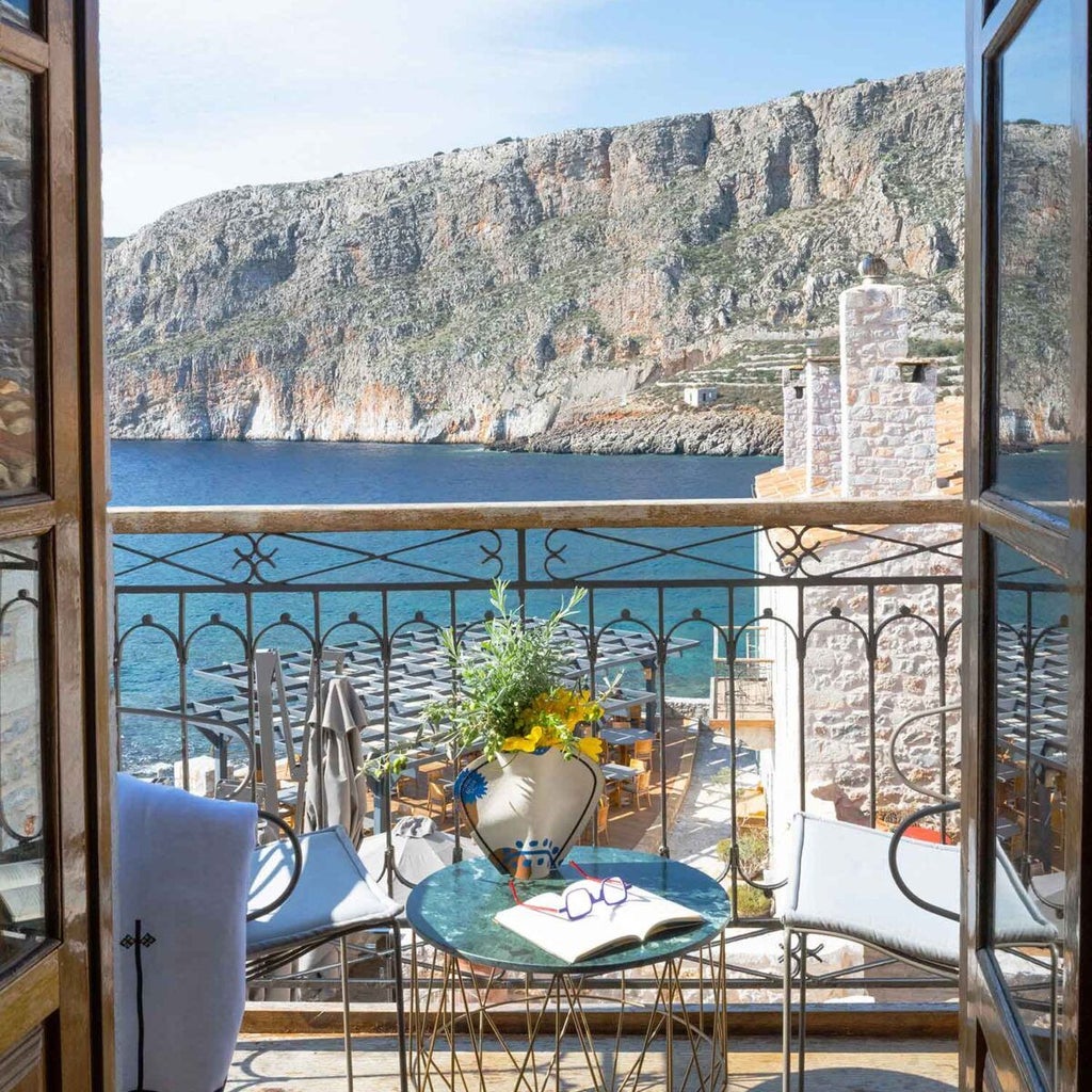 Luxurious stone-built seaside hotel with traditional Greek architecture, nestled against azure Aegean waters, featuring elegant stone walls and rustic wooden balconies