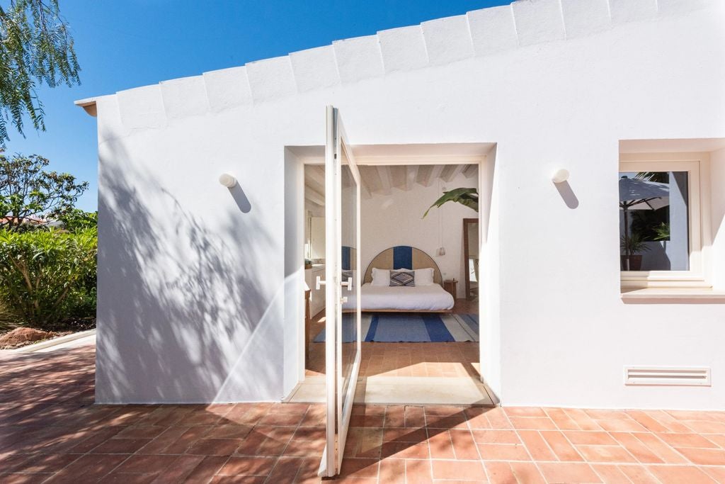 Bright, minimalist hotel room with whitewashed walls, rustic wooden beams, plush queen bed, and natural rattan accents overlooking a terrace