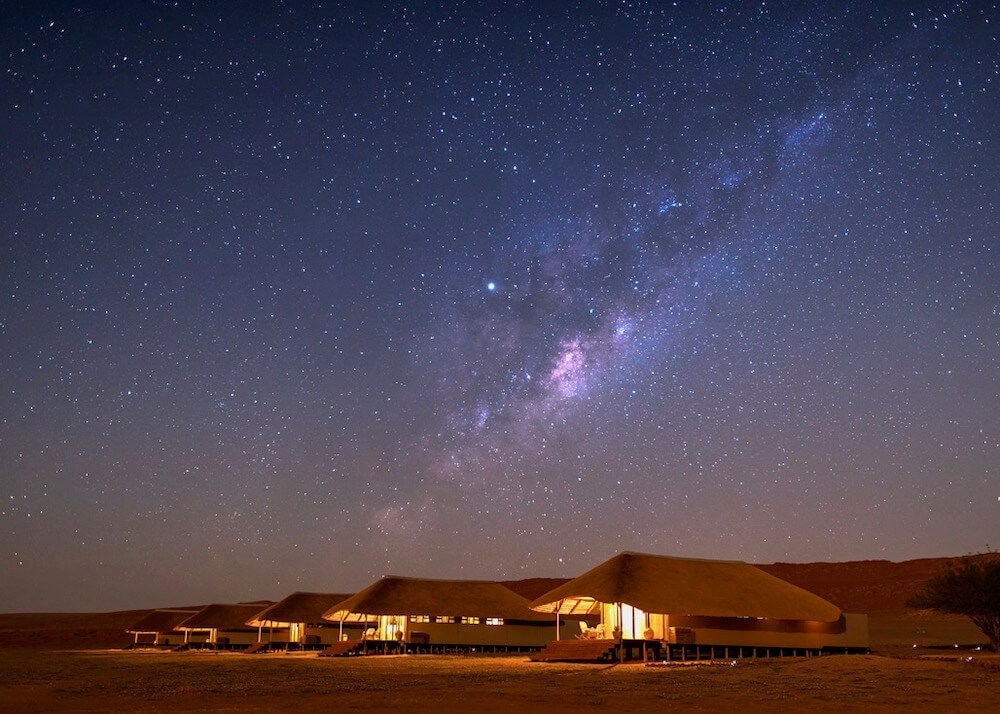 Luxurious desert lodge with thatched-roof cottage overlooking vast red sand dunes of NamibRand Nature Reserve at sunset