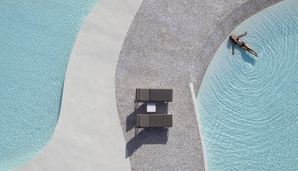 Elegant white hotel buildings with private infinity pools nestled into a hillside, overlooking the Mediterranean Sea at sunset