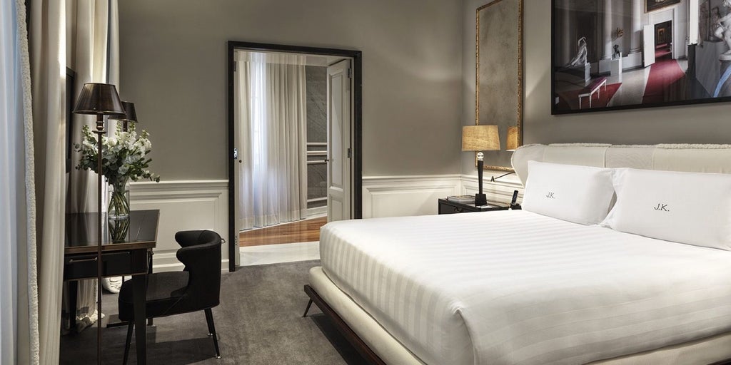 Elegant grand deluxe hotel room with plush cream furnishings, soft lighting, and sophisticated Italian design overlooking historic Rome's architectural landscape