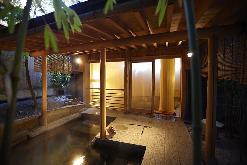 Traditional Japanese ryokan with wooden architecture features private outdoor onsen bath surrounded by zen garden and lantern lighting