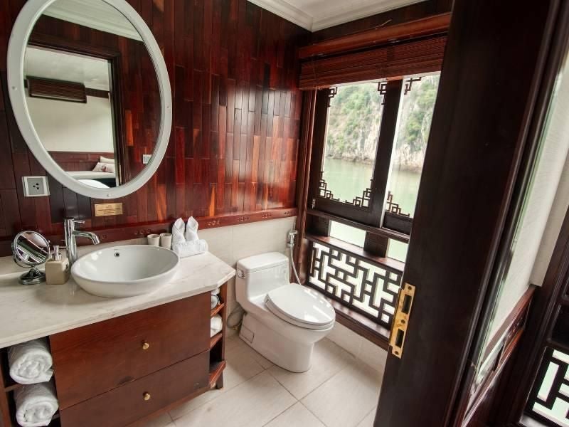 Luxurious Bhaya Cruises deluxe cabin with elegant wood paneling, plush white bedding, and panoramic windows overlooking emerald Halong Bay waters