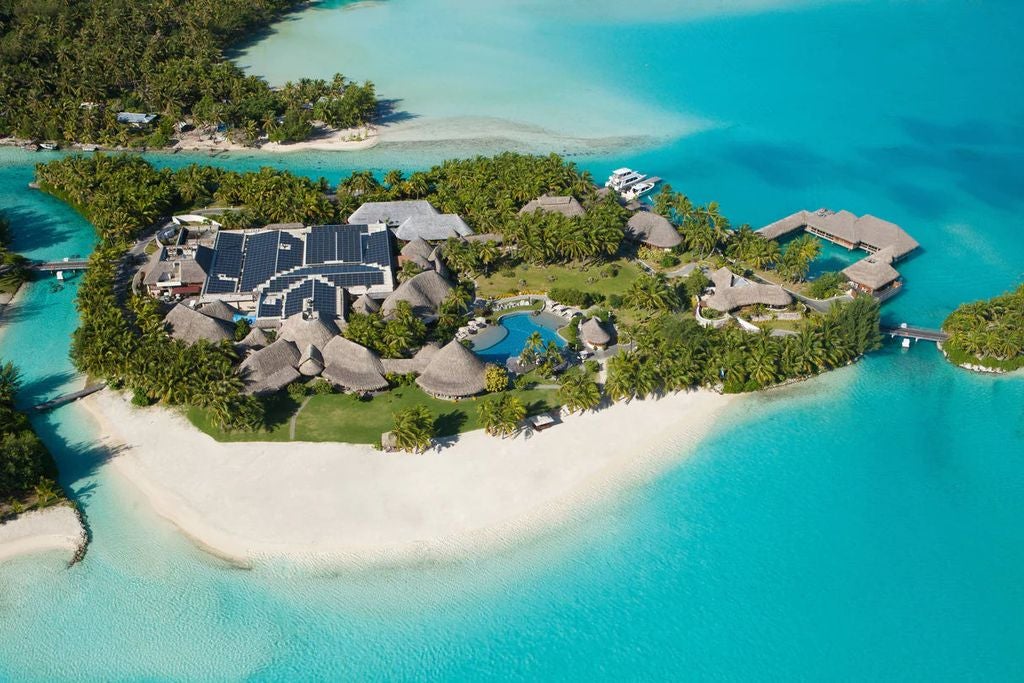 Overwater luxury bungalows connected by wooden walkways stretch into turquoise lagoon, backed by lush mountains and coral reefs