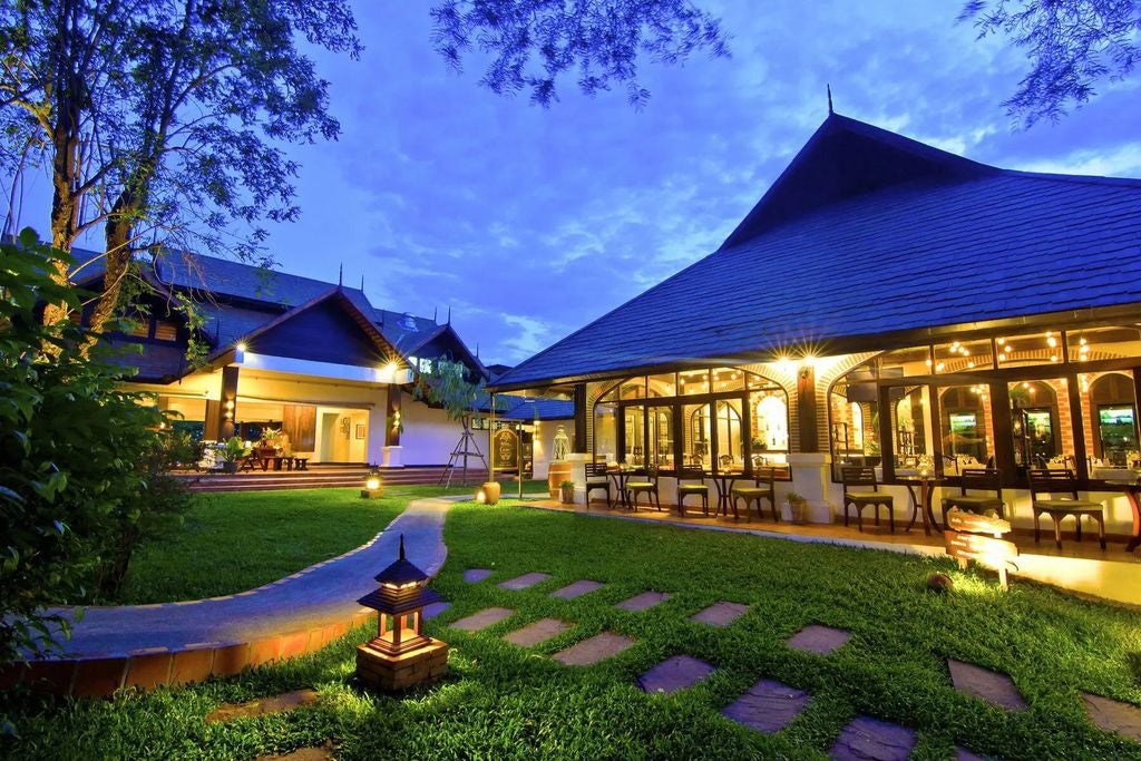 Elegant Thai hotel exterior with illuminated white facade, grand entrance columns, tropical gardens and reflection pool at dusk