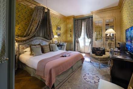 Elegant Provençal-style deluxe hotel room with lavish white furnishings, soft blue accents, and ornate chandelier in a refined French boutique hotel setting