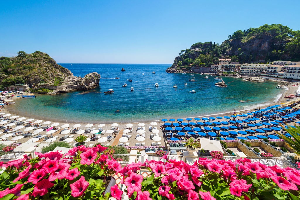 Elegant Mediterranean hotel with white terraces overlooking turquoise Taormina Bay, flanked by palm trees and lush gardens