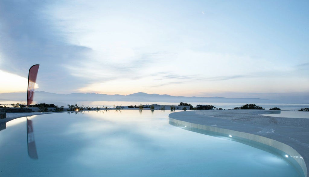 Elegant Mediterranean hotel with infinity pool overlooking Aegean Sea, white-washed buildings, and palm trees at sunset in Greece