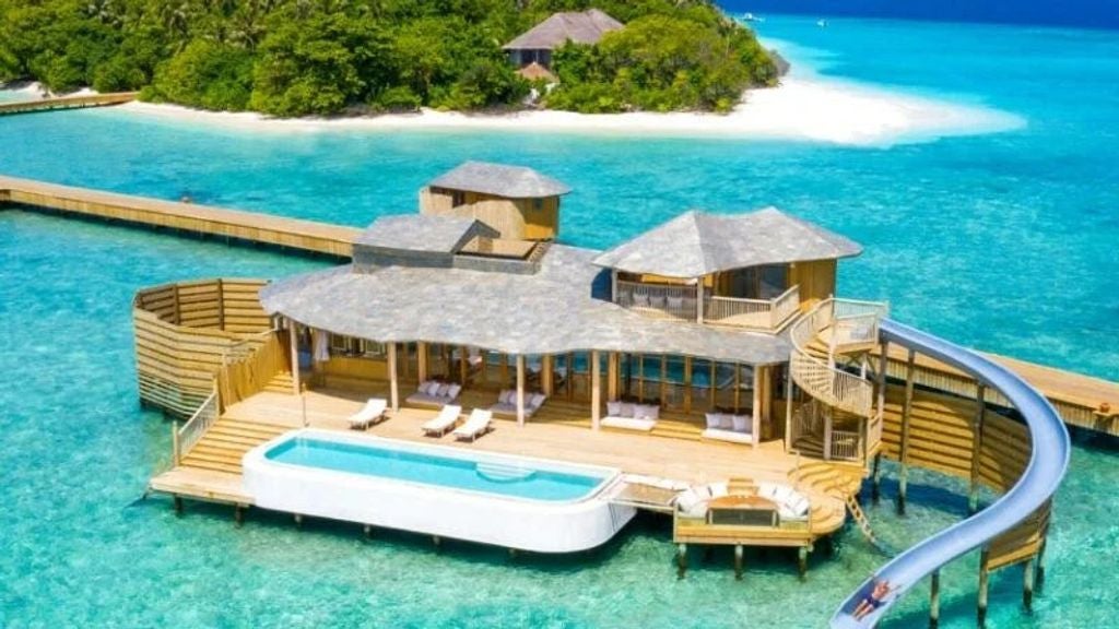 Luxurious overwater villa with wooden walkway stretches over crystal-clear turquoise Maldivian lagoon beneath sunny blue skies