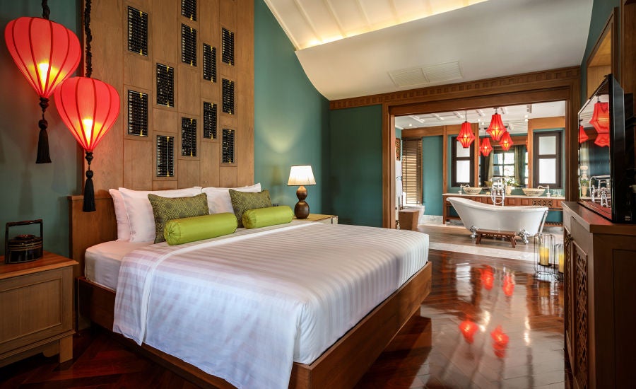 Elegant colonial-style suite with four-poster bed, ornate Chinese furniture, dark wood accents, and floor-to-ceiling windows overlooking tropical gardens