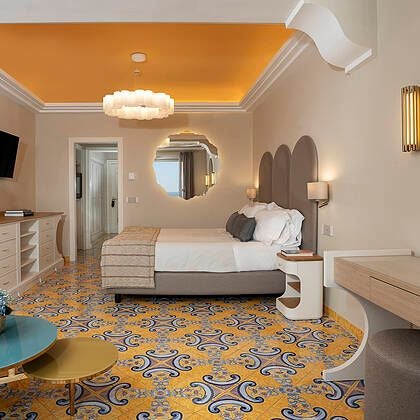 Elegant Italian hotel room with plush king bed, ornate gold-framed mirror, marble bathroom, and panoramic view of scenic coastal landscape