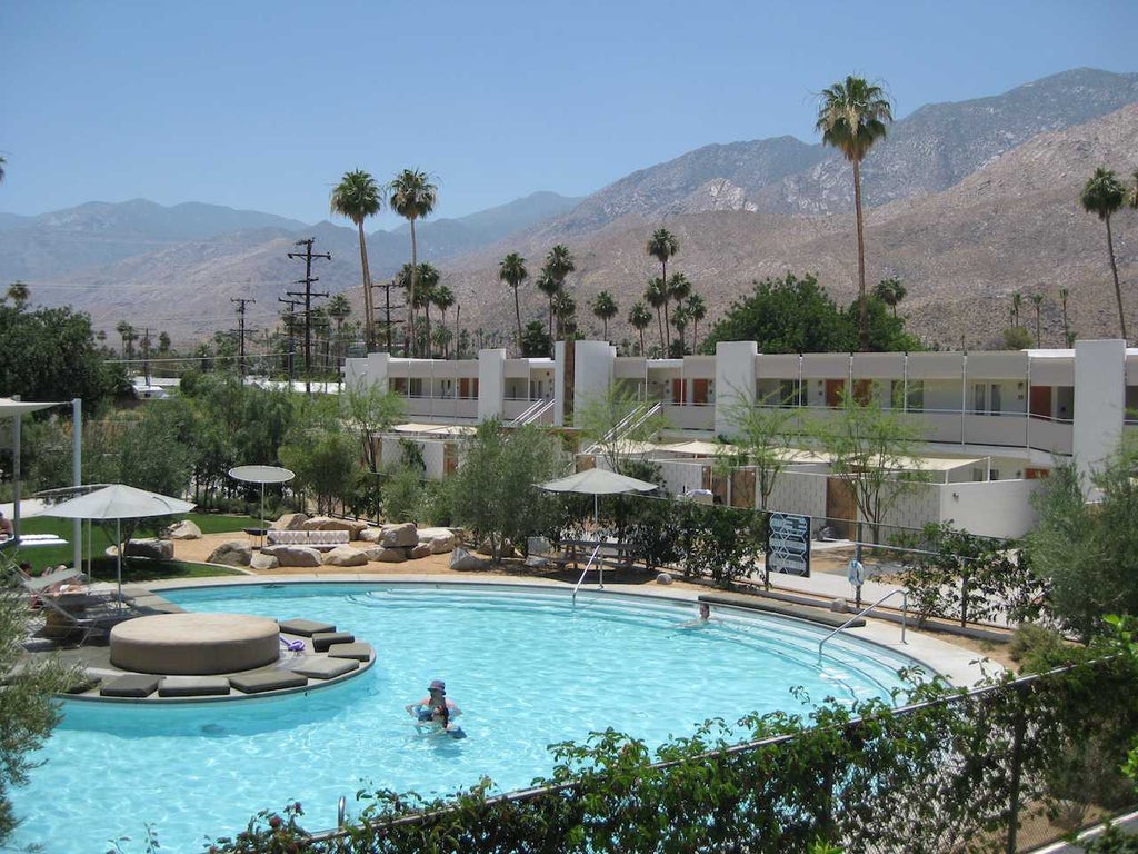 Mid-century modern desert hotel with sleek white exterior, palm trees, and chic minimalist pool area set against dramatic mountain backdrop