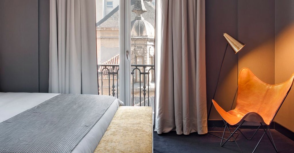 Elegant deluxe hotel room with floor-to-ceiling windows overlooking Madrid's cityscape, featuring modern decor, plush bedding, and soft ambient lighting.