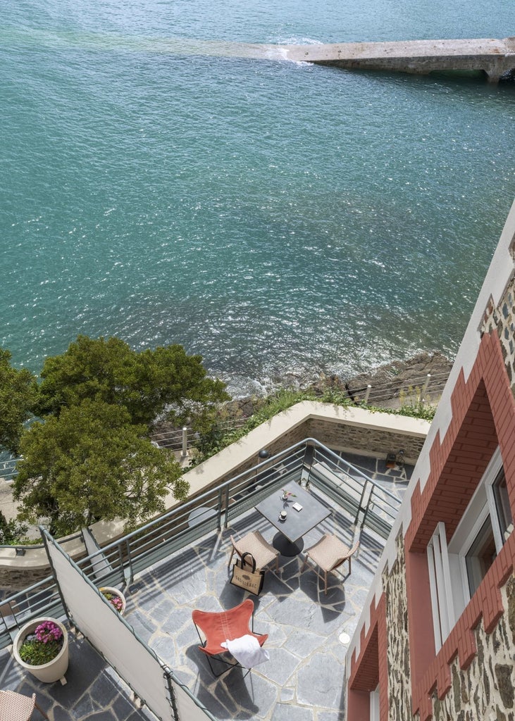 Luxurious seaside hotel nestled on rocky coastline, elegant blue-gray facade with white trim, overlooking dramatic Brittany cliffs and azure waters