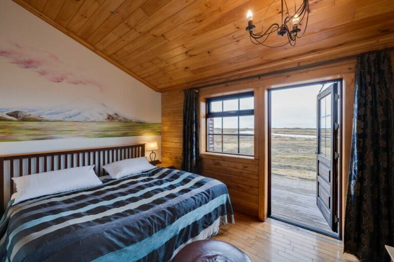 Spacious deluxe suite with wooden accents, plush king bed, rustic chandelier, and large windows showcasing Icelandic countryside views