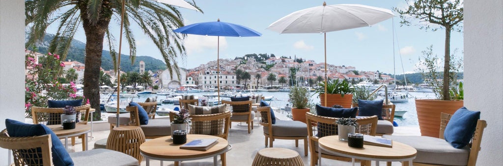 Modern luxury waterfront hotel with white Mediterranean facade, rooftop infinity pool, and palm-lined terrace overlooking Hvar marina at sunset