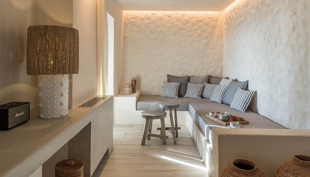 Elegant white-washed hotel room with minimalist Cycladic design, featuring crisp linens, blue accent pillows, and panoramic Aegean Sea view from large window