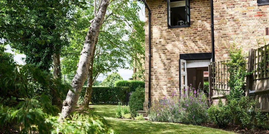 Luxurious East Cottage Suite at Coworth Park, featuring elegant country-style decor with plush white bedding, antique furniture, and large windows overlooking manicured gardens.