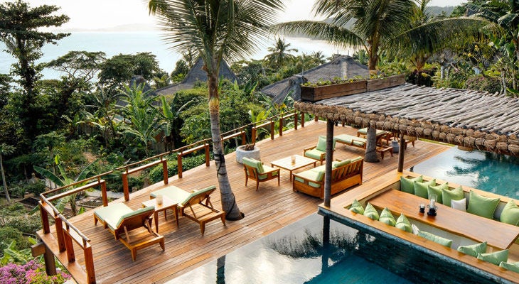 Luxurious thatched-roof villa on stilts overlooking pristine Indonesian beach with infinity pool merging into ocean horizon at sunset