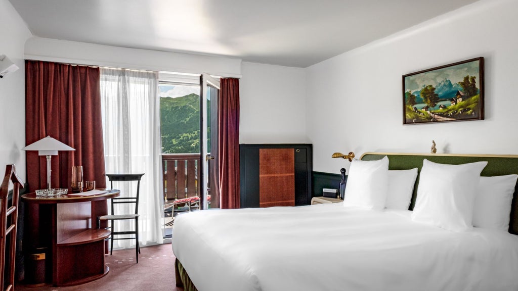 Modern Swiss alpine junior suite with minimalist wood paneling, plush white bedding, and floor-to-ceiling windows revealing stunning mountain landscape in soft evening light