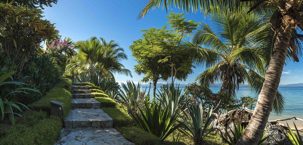 Luxurious wooden bungalow perched on tropical hillside overlooking turquoise ocean bay, surrounded by lush palm trees and native flora