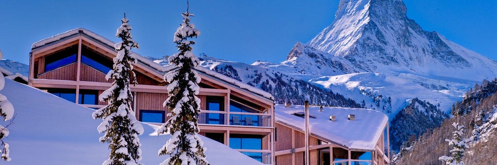 Modern mountain lodge with panoramic glass windows reflecting the snow-capped Matterhorn peak against a clear Swiss Alpine sky