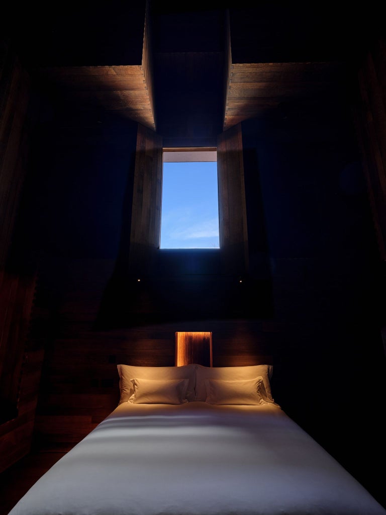 Minimalist luxury hotel room with sculptural wooden bed, stark white walls, and angular design by renowned artist, illuminated by soft ambient lighting