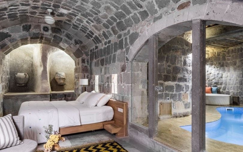 Elegant stone-walled suite with traditional Turkish decor, plush white bedding, arched windows overlooking Cappadocia's unique rocky landscape at sunset