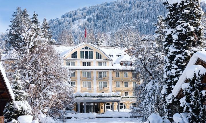 Opulent Swiss alpine hotel with grand white facade, ornate windows, and manicured gardens against a backdrop of snow-capped mountain peaks at sunset