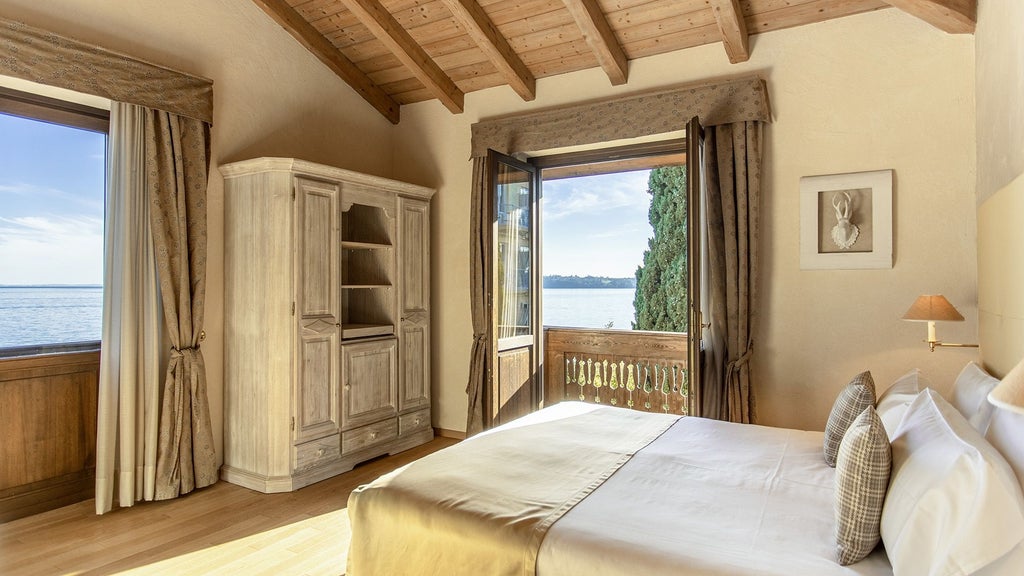 Elegant hotel room with partial Lake Garda views, featuring marble floors, cream walls, chic furnishings and a king-sized bed with crisp linens