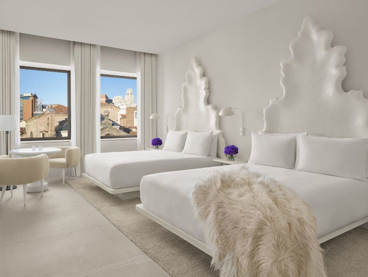 Modern hotel room with two queen beds, city views through floor-to-ceiling windows, wood accents, and designer pendant lighting