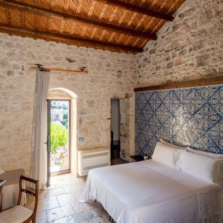 Elegant Mediterranean-style hotel room with stone walls, rustic wooden furniture, soft white linens, and warm natural light streaming through arched windows