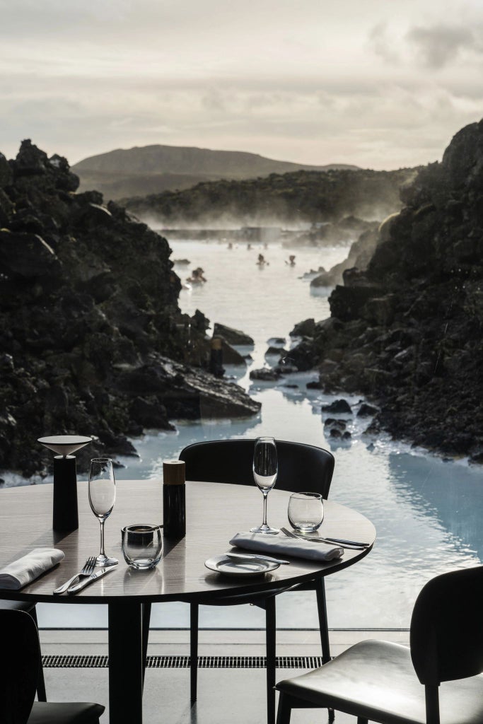 Minimalist Icelandic hotel suite with floor-to-ceiling windows overlooking a tranquil blue lagoon, featuring sleek Nordic design and natural stone elements